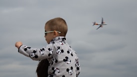 A young child wearing a Mickey Mouse patterned jacket and sunglasses is sitting on someone's shoulders. In the background, an airplane is seen flying across a cloudy sky.