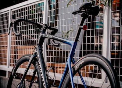 A sleek, modern road bike is positioned against a metal fence. The frame is primarily black with blue accents, and the branding 'Specialized' is visible. The bike features drop handlebars and high-performance wheels.