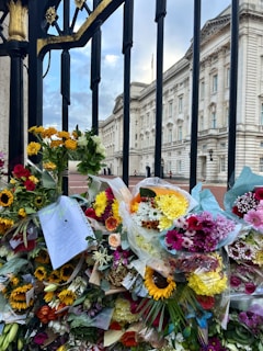 Beautiful bouquets of colorful flowers are laid against the ornate black and gold metal gate of a large, grand building. The flowers include sunflowers, roses, chrysanthemums, and lilies in vibrant hues of yellow, pink, red, and white, creating a striking display. A note is visible among the flowers, adding a personal touch.