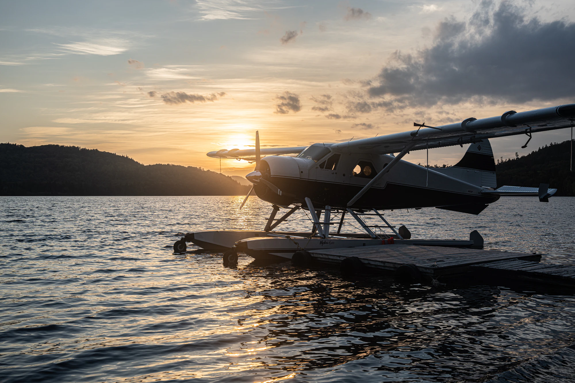 Float plane tied at a dock on a mirrored lake at dusk with dark mountain silhouettes beyond