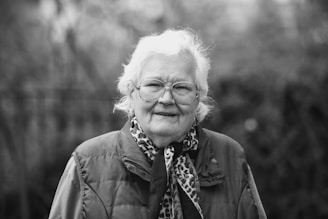 An elderly person with white hair and glasses is wearing a patterned scarf and a padded jacket. The background is blurred, suggesting an outdoor setting.