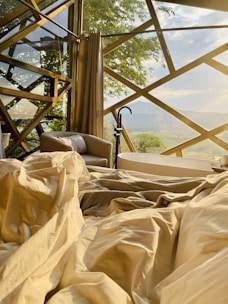 A cozy interior space with an unmade bed featuring soft, cream-colored sheets. In the background, large windows with a geometric wooden frame provide a view of lush greenery and a distant landscape. A plush armchair with a striped cushion is positioned near a freestanding bathtub facing the window, creating a serene, inviting atmosphere bathed in warm, natural light.