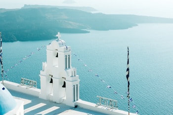 A white church with bells stands in the foreground, overlooking a vast expanse of turquoise sea and distant islands. Blue and white flags are draped across the building, consistent with a Greek theme. The architecture is characterized by classic Greek elements, with clear skies and gentle sunlight. Mountains can be seen across the water, giving a serene and peaceful ambiance.