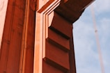 A close-up view of a structure made of orange-painted steel beams and rivets, likely part of a bridge. The design includes geometric shapes with distinct lines and shadows casting depth. A segment of supporting cables is visible against a blue sky in the background.