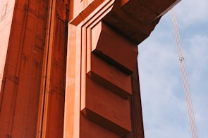 A close-up view of a structure made of orange-painted steel beams and rivets, likely part of a bridge. The design includes geometric shapes with distinct lines and shadows casting depth. A segment of supporting cables is visible against a blue sky in the background.
