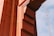 A close-up view of a structure made of orange-painted steel beams and rivets, likely part of a bridge. The design includes geometric shapes with distinct lines and shadows casting depth. A segment of supporting cables is visible against a blue sky in the background.