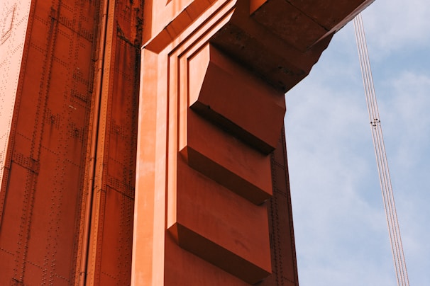 A close-up view of a structure made of orange-painted steel beams and rivets, likely part of a bridge. The design includes geometric shapes with distinct lines and shadows casting depth. A segment of supporting cables is visible against a blue sky in the background.