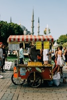 A colorful street food cart offers roasted corn for sale. The cart has a red design with a striped canopy and displays signs showing prices in Turkish Lira. Corn is neatly arranged on the cart, ready for customers. People walk by, and a historical mosque with tall minarets is visible in the background, suggesting a bustling urban setting.