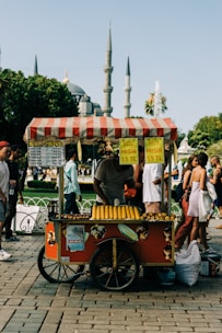 A colorful street food cart offers roasted corn for sale. The cart has a red design with a striped canopy and displays signs showing prices in Turkish Lira. Corn is neatly arranged on the cart, ready for customers. People walk by, and a historical mosque with tall minarets is visible in the background, suggesting a bustling urban setting.