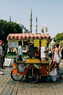A colorful street food cart offers roasted corn for sale. The cart has a red design with a striped canopy and displays signs showing prices in Turkish Lira. Corn is neatly arranged on the cart, ready for customers. People walk by, and a historical mosque with tall minarets is visible in the background, suggesting a bustling urban setting.