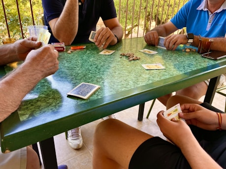 Four people are gathered around a green-speckled table, engaged in a card game. The players are holding cards and there are playing cards and coins scattered across the table. A glass with a drink, a pack of cigarettes, and other small items are visible on the table.