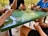 Four people are gathered around a green-speckled table, engaged in a card game. The players are holding cards and there are playing cards and coins scattered across the table. A glass with a drink, a pack of cigarettes, and other small items are visible on the table.