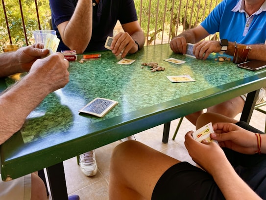 Four people are gathered around a green-speckled table, engaged in a card game. The players are holding cards and there are playing cards and coins scattered across the table. A glass with a drink, a pack of cigarettes, and other small items are visible on the table.