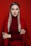 A woman with long, straight, blonde hair poses against a solid red background. She wears a red turtleneck and matching red jacket, with her arms crossed confidently. Her makeup is professionally done, with a focus on dark eyeliner and subtle lipstick.