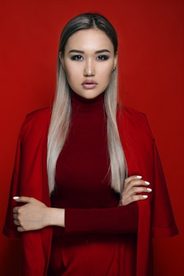 A woman with long, straight, blonde hair poses against a solid red background. She wears a red turtleneck and matching red jacket, with her arms crossed confidently. Her makeup is professionally done, with a focus on dark eyeliner and subtle lipstick.