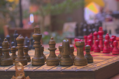 A chess board set up with wooden pieces, featuring a foreground of dark brown and red chess pieces on a light-colored board. The focus is on the chess pieces, showing intricate details and texture. The background is blurred, with hints of greenery and a soft, ambient setting.