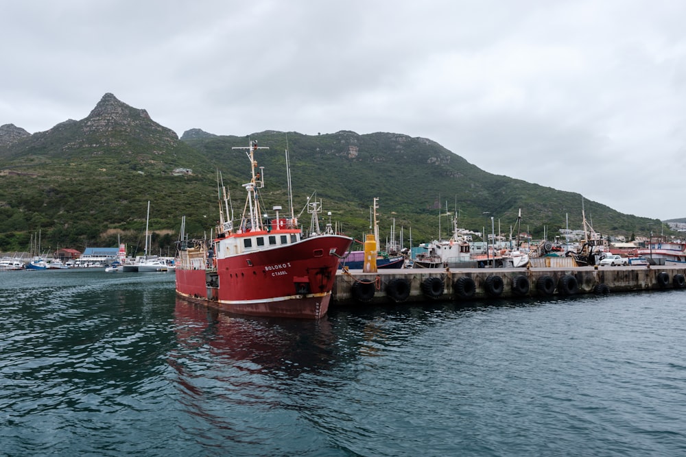 a red and white boat in the water by a dock with mountains in the background