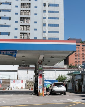 A gas station with a car refueling under a canopy. The station is situated in an urban area, surrounded by tall buildings. A person is standing next to the vehicle, and signage is visible on the station's pillars. Traffic cones are placed nearby, and the background includes a blue sky and a mix of residential and commercial buildings.