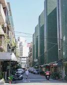 An urban alleyway flanked by tall buildings on both sides. The right side is dominated by a series of green-colored buildings, while the left side has concrete structures with balconies and plants. Several vehicles, including scooters and cars, are parked along the sides of the street. A few people walk at the far end of the alley. The sky is clear and blue.
