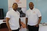 Two men wearing white polo shirts with a logo stand in front of a banner. The banner displays the name 'Inkanyeti Foundation' and an image of a person with glasses. There is a table with items in the background to the right.
