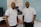 Two men wearing white polo shirts with a logo stand in front of a banner. The banner displays the name 'Inkanyeti Foundation' and an image of a person with glasses. There is a table with items in the background to the right.