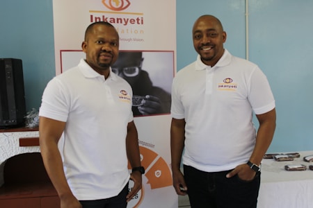 Two men wearing white polo shirts with a logo stand in front of a banner. The banner displays the name 'Inkanyeti Foundation' and an image of a person with glasses. There is a table with items in the background to the right.