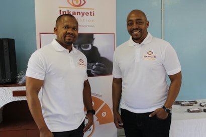 Two men wearing white polo shirts with a logo stand in front of a banner. The banner displays the name 'Inkanyeti Foundation' and an image of a person with glasses. There is a table with items in the background to the right.