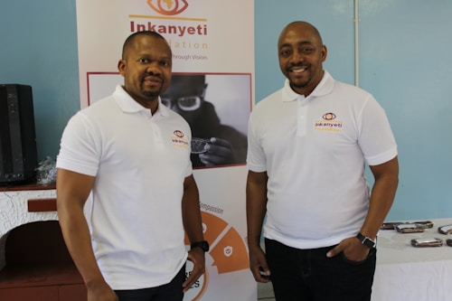Two men wearing white polo shirts with a logo stand in front of a banner. The banner displays the name 'Inkanyeti Foundation' and an image of a person with glasses. There is a table with items in the background to the right.