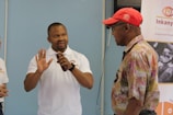 A man in a white polo shirt is gesturing with his hands while speaking to an older man wearing a red cap and a floral shirt. A partially visible person stands to the side. A banner from the Inkanyezi Foundation is displayed in the background.