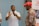 A man in a white polo shirt is gesturing with his hands while speaking to an older man wearing a red cap and a floral shirt. A partially visible person stands to the side. A banner from the Inkanyezi Foundation is displayed in the background.