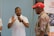 A man in a white polo shirt is gesturing with his hands while speaking to an older man wearing a red cap and a floral shirt. A partially visible person stands to the side. A banner from the Inkanyezi Foundation is displayed in the background.