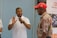 A man in a white polo shirt is gesturing with his hands while speaking to an older man wearing a red cap and a floral shirt. A partially visible person stands to the side. A banner from the Inkanyezi Foundation is displayed in the background.
