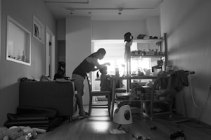 A monochrome photo depicting a cluttered room with a person tending to a child at a table. The room is filled with scattered items, giving a lived-in feel. Shelves in the background hold various objects, and light is streaming in from a window, casting shadows on the floor.