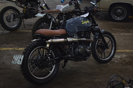 Several motorcycles, including a prominently featured custom Suzuki with a brown leather seat, are parked indoors. The environment appears to be a well-lit garage or workshop with a concrete floor.