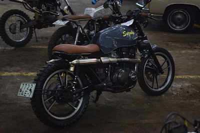 Several motorcycles, including a prominently featured custom Suzuki with a brown leather seat, are parked indoors. The environment appears to be a well-lit garage or workshop with a concrete floor.