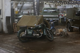 A vintage motorcycle and a blue scooter are parked inside a dimly lit garage. There's a canvas tent set up nearby. Various cars are visible in the background, alongside string lights hanging from the ceiling. A bundle of sticks is arranged on the ground, simulating a campfire.