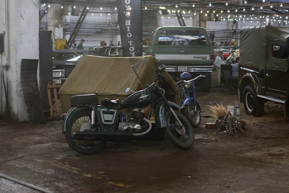 A vintage motorcycle and a blue scooter are parked inside a dimly lit garage. There's a canvas tent set up nearby. Various cars are visible in the background, alongside string lights hanging from the ceiling. A bundle of sticks is arranged on the ground, simulating a campfire.