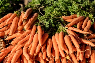 A large pile of fresh carrots with green tops, arranged closely together. The carrots vary in size and have a vibrant orange color, complemented by the green leaves, creating a natural and earthy look.