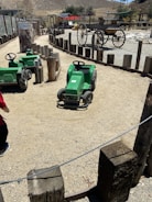 A dusty path with green pedal tractors surrounded by wooden posts. In the background, there is an old wooden wagon and a small building with a red roof. The area is set in a dry, hilly landscape.