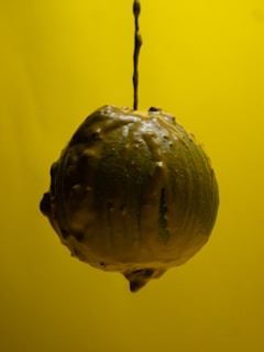 A sphere covered with a glossy, textured coating is suspended against a vibrant yellow background. The surface appears to be dripping with a creamy substance, suggesting motion or a recent application of the material.