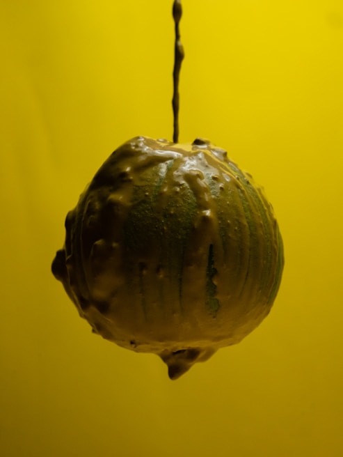A sphere covered with a glossy, textured coating is suspended against a vibrant yellow background. The surface appears to be dripping with a creamy substance, suggesting motion or a recent application of the material.