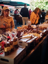 A bustling market scene where various confectionery items are displayed on a counter, including chocolates and wrapped goods, drawing the attention of shoppers. People are casually dressed, with a woman in a beige trench coat holding a can and another woman in an orange jacket browsing the stalls. The market is lively, with a green canopy overhead and several customers in the background.