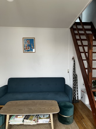 A cozy living space features a dark green sofa with a wooden coffee table in front of it. Several magazines are stored underneath the table. A small, round, matching green ottoman is placed near the sofa. On the wall, there is a colorful framed artwork depicting people by the water. A wooden staircase with a modern design is located on the right, leading upward. The floor is wooden, adding warmth to the room. Overall, the area appears organized and inviting.