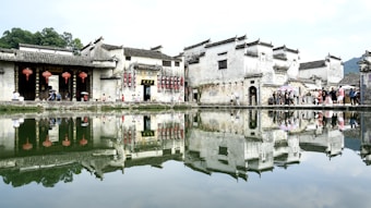 Ancient Chinese buildings with traditional architecture are reflected in a calm water body. Red lanterns hang from the eaves, and a group of people walk along the path by the water, some carrying umbrellas. The scene is completed by distant greenery and a partly cloudy sky.