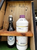 Two bottles are placed on a small wooden shelf. On the left is a dark glass bottle with a white flip-top cap, labeled 'Das Echte Schwaben Bräu Märzen' with an illustration of a bearded man. On the right is a plastic bottle with a purple cap, labeled 'ATEV Gruppe KühlerSchutz A12 Plus', and hand-labeled 'VW-Bus'. Above each bottle are handwritten labels 'Fahrer' and 'Auto', with arrows pointing to each.