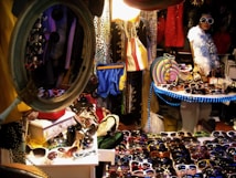 A cluttered market stall brimming with a variety of colorful sunglasses displayed on tables under bright lights. A mannequin dressed in vibrant, trendy clothing and wearing oversized sunglasses stands to the right, surrounded by eclectic fashion items including hats, bags, and a feather boa. The setup is packed with different patterns and textures, surrounded by racks and mirrors, creating a lively, vibrant atmosphere.