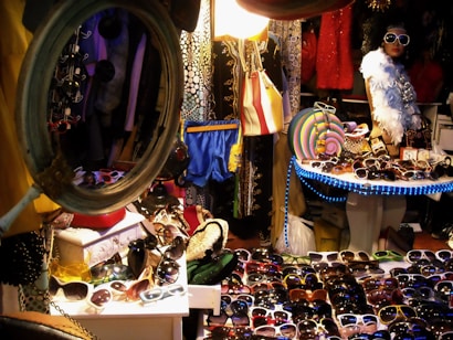 A cluttered market stall brimming with a variety of colorful sunglasses displayed on tables under bright lights. A mannequin dressed in vibrant, trendy clothing and wearing oversized sunglasses stands to the right, surrounded by eclectic fashion items including hats, bags, and a feather boa. The setup is packed with different patterns and textures, surrounded by racks and mirrors, creating a lively, vibrant atmosphere.