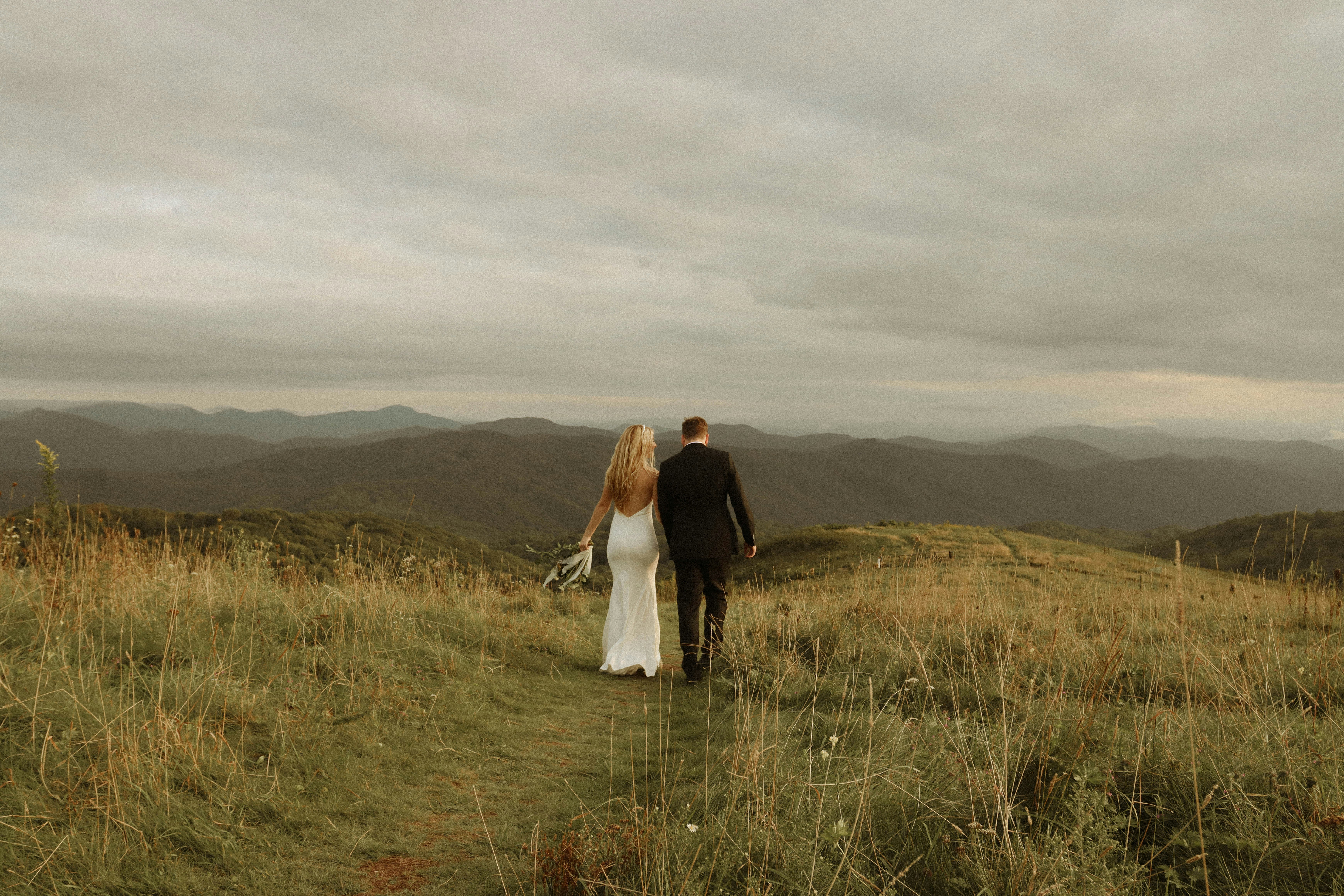 a man and woman standing in a field of tall grass with hills in the background