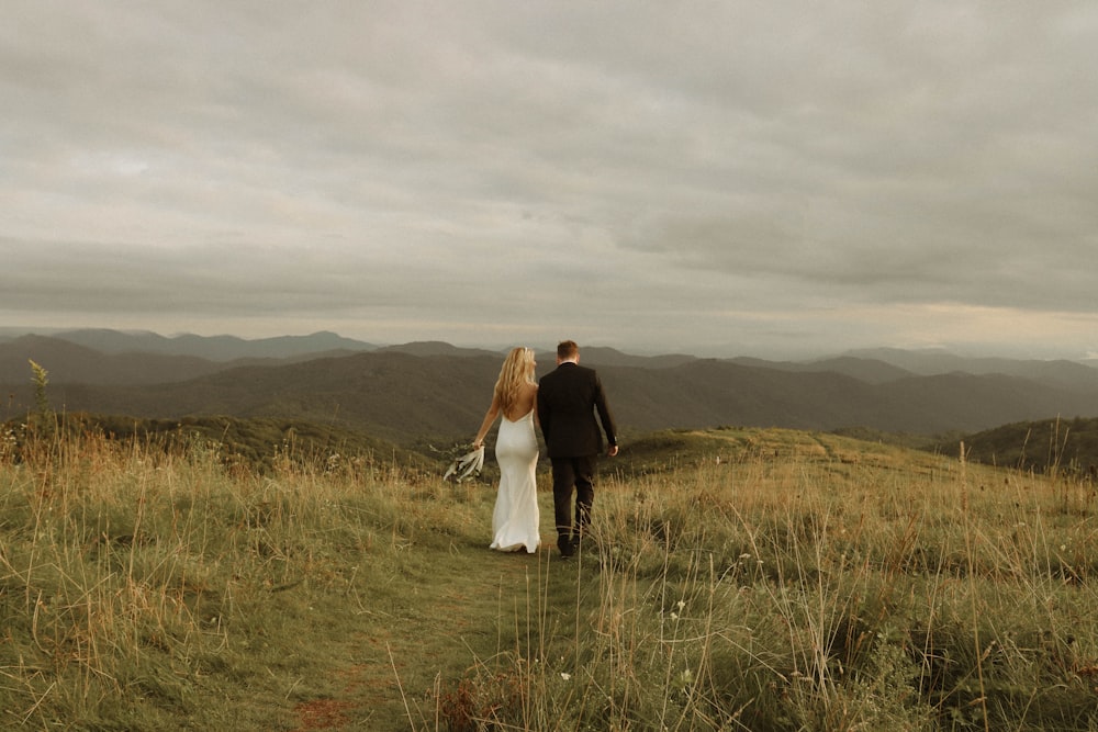 a man and woman standing in a field of tall grass with hills in the background