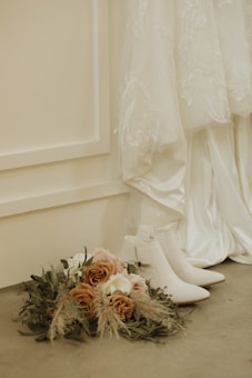 A bouquet of flowers is placed on a gray floor next to a pair of white ankle boots. In the background, there is a detailed white wedding dress with intricate lace and floral patterns, hanging against a beige wall with panel designs.